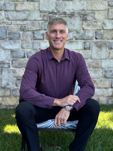 Smiling person in a purple shirt and black pants sitting on a chair outdoors by a stone wall