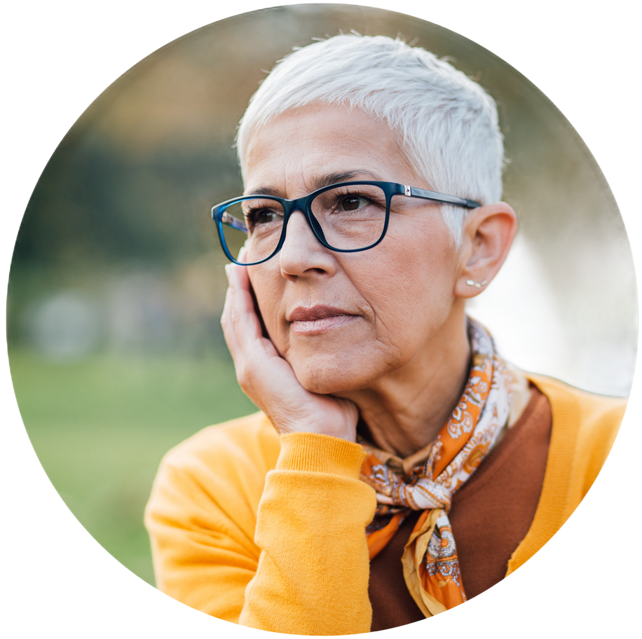 Portrait of a person with short white hair, glasses, and a yellow sweater, resting their cheek on their hand outdoors
