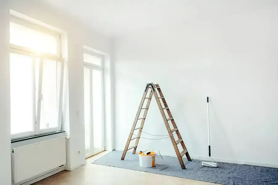 Bright white room under renovation with a stepladder, paint bucket, and drop cloth by the windows