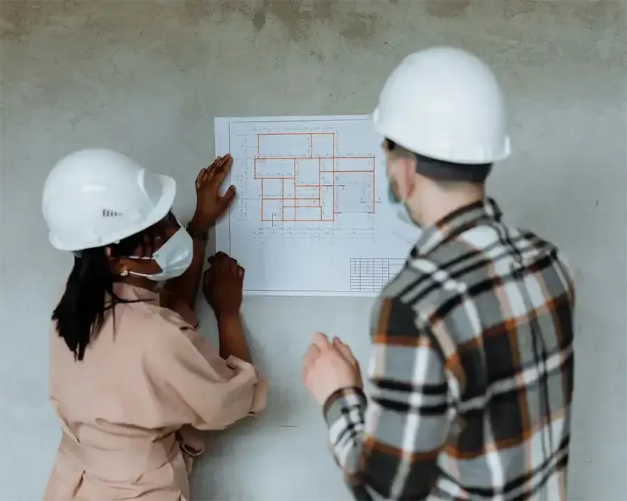 Two construction workers in white hard hats review blueprints on a wall at a job site.