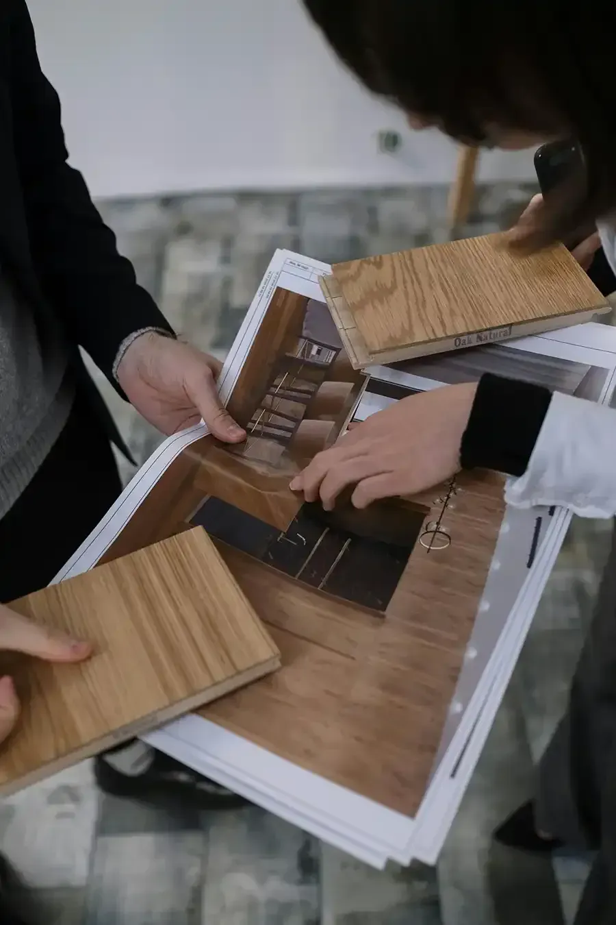 Hands assembling a wooden model house over papers and wood samples on a table