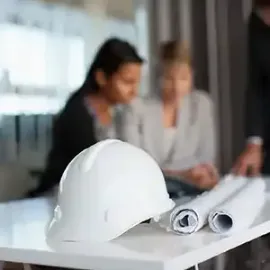 White hard hat and rolled blueprints on a table with two people blurred in the background