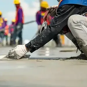 Worker smoothing wet concrete with a trowel at a construction site, with blurred workers in the background