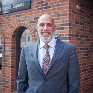 A man in a suit and tie stands in front of a brick building that says public square