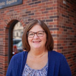 A woman wearing glasses and a blue sweater is smiling in front of a brick building.