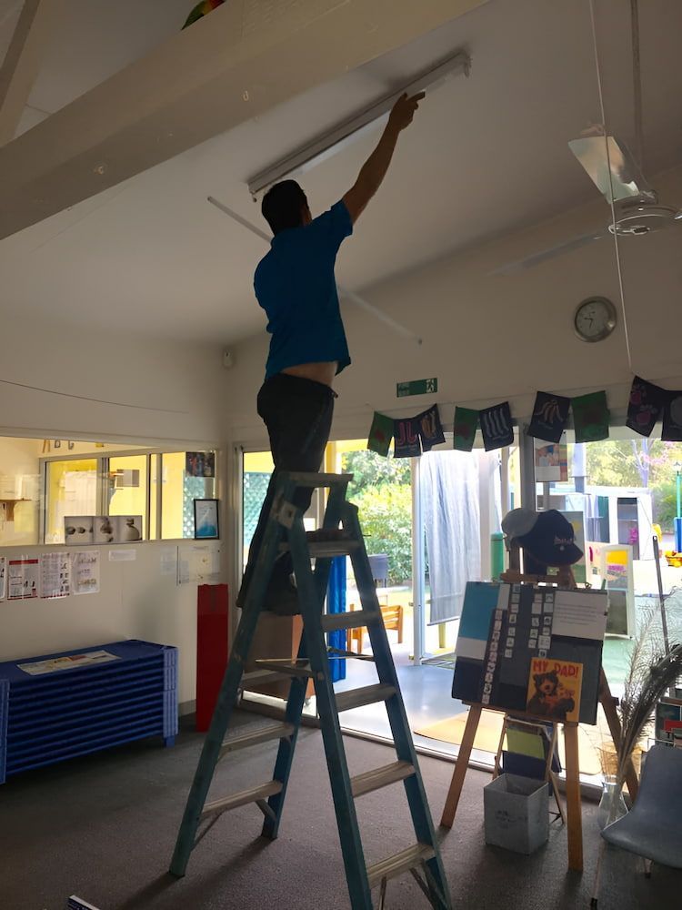 A Man Is Standing On A Ladder Working On A Light Fixture — Levi's Electrics in Nerimbera, QLD