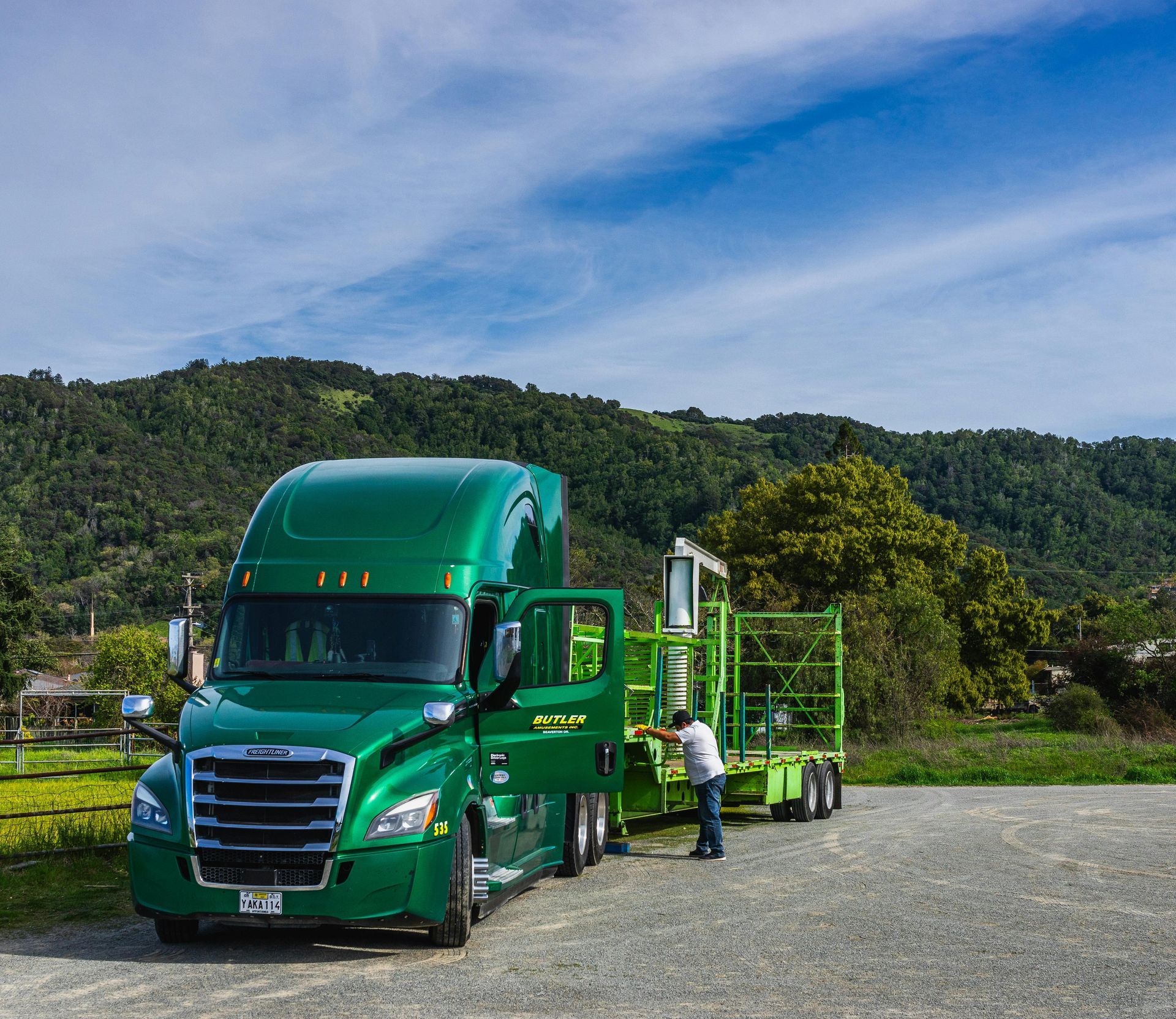 Green semi-truck parked on a gravel lot with a person standing beside it, hills and trees in the background