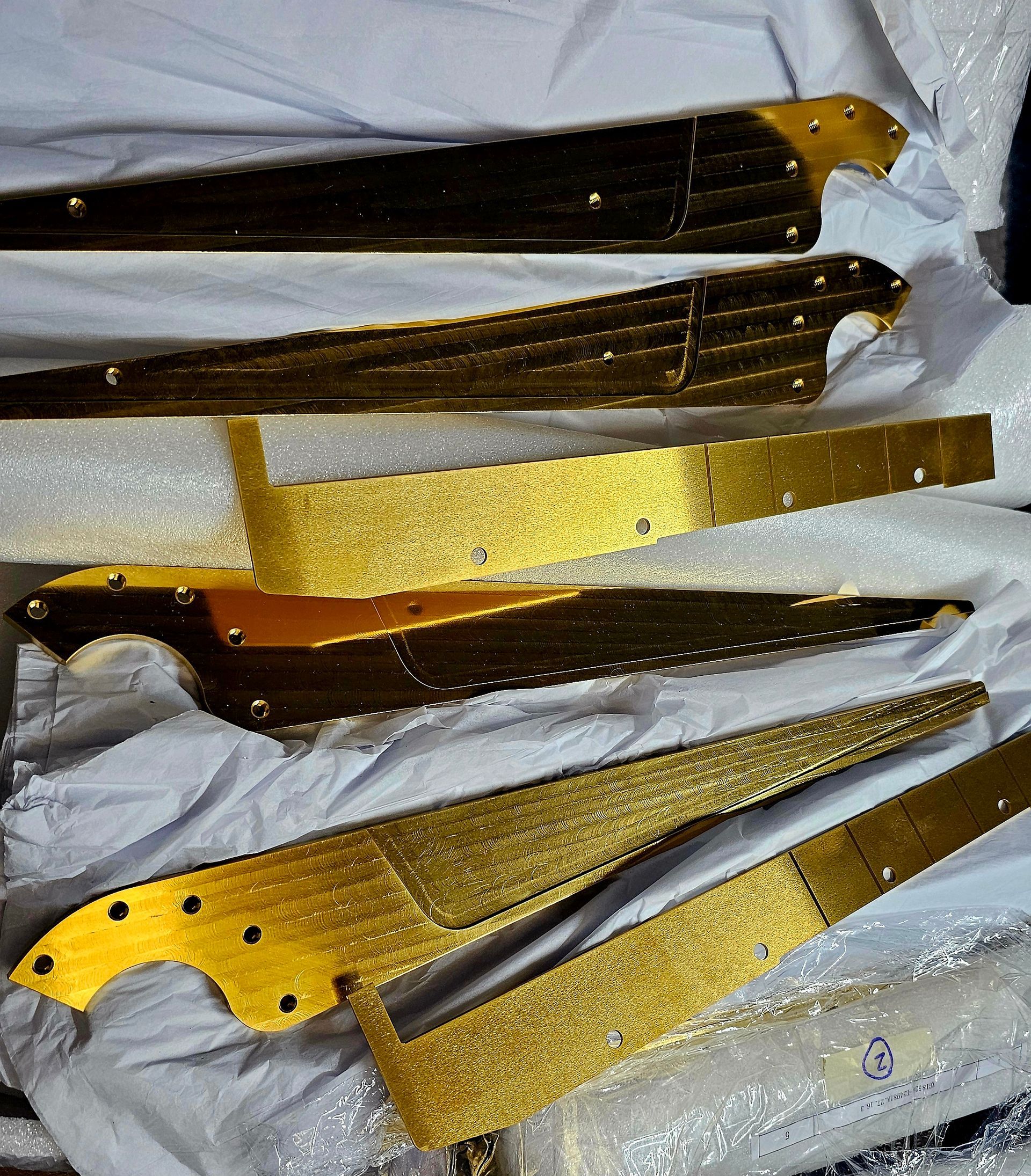 Assorted gold-and-black decorative knife-shaped objects with textured handles on a white cloth background