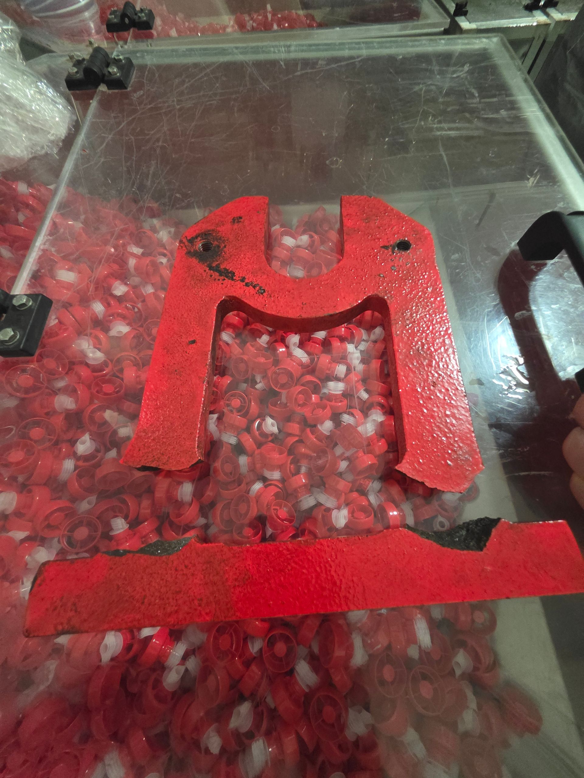 Red plastic U-shaped bracket with a small hole, resting on a clear surface over red pebbles.