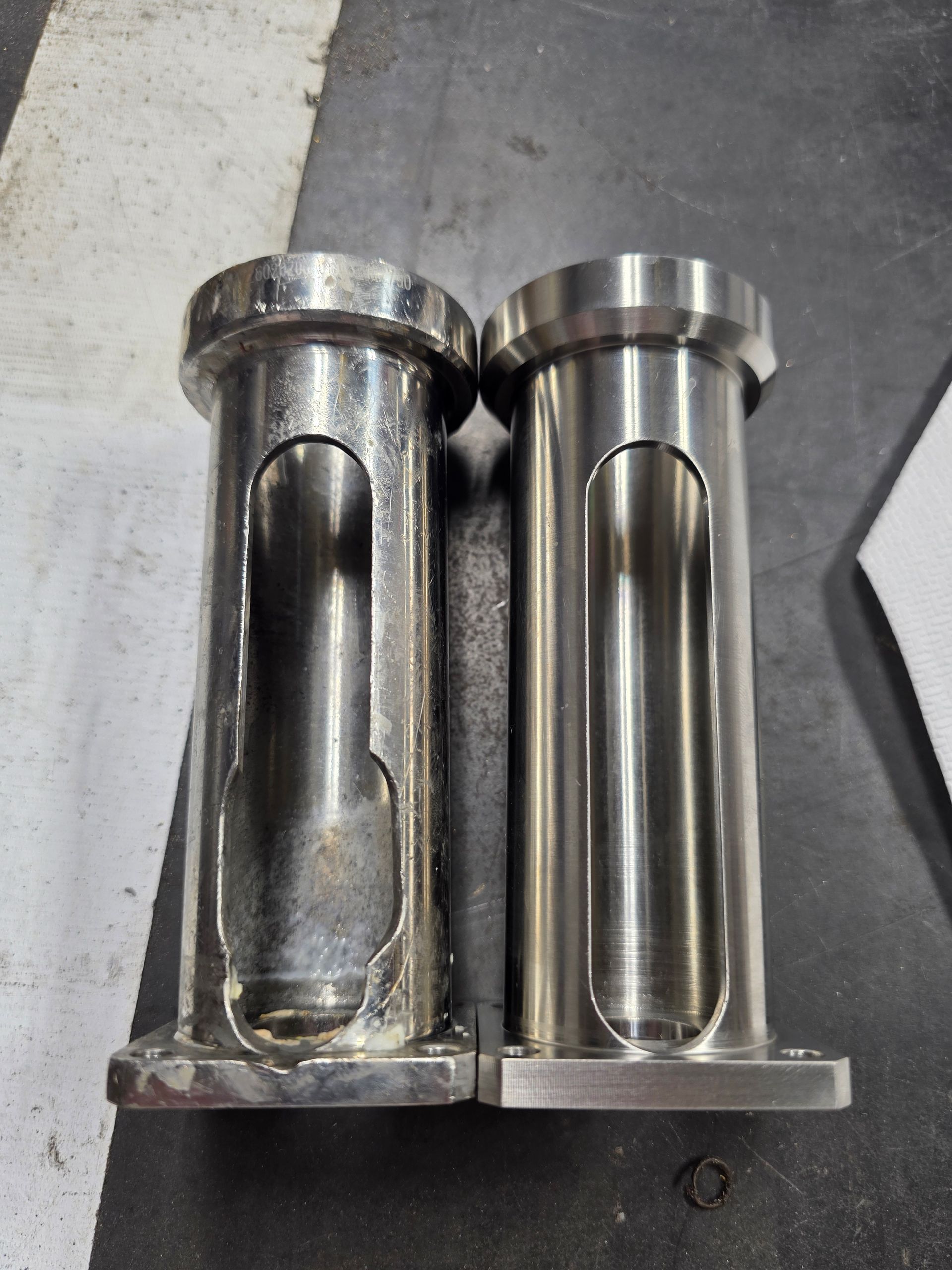 Two metal cylindrical parts on a gray surface, one dark and worn, one shiny and polished.