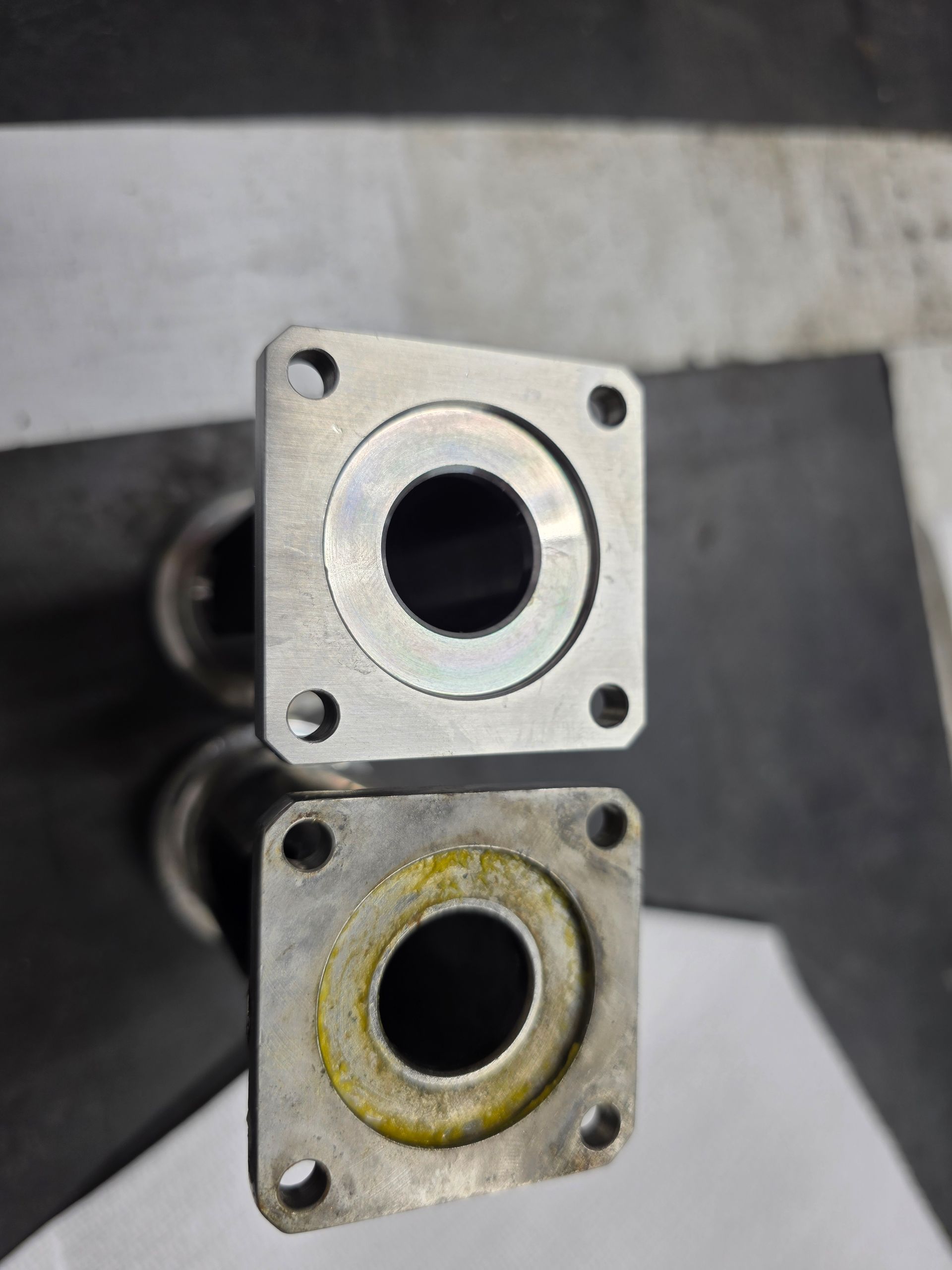 Two square metal flanges with central holes, one silver and one rust-stained, stacked vertically.