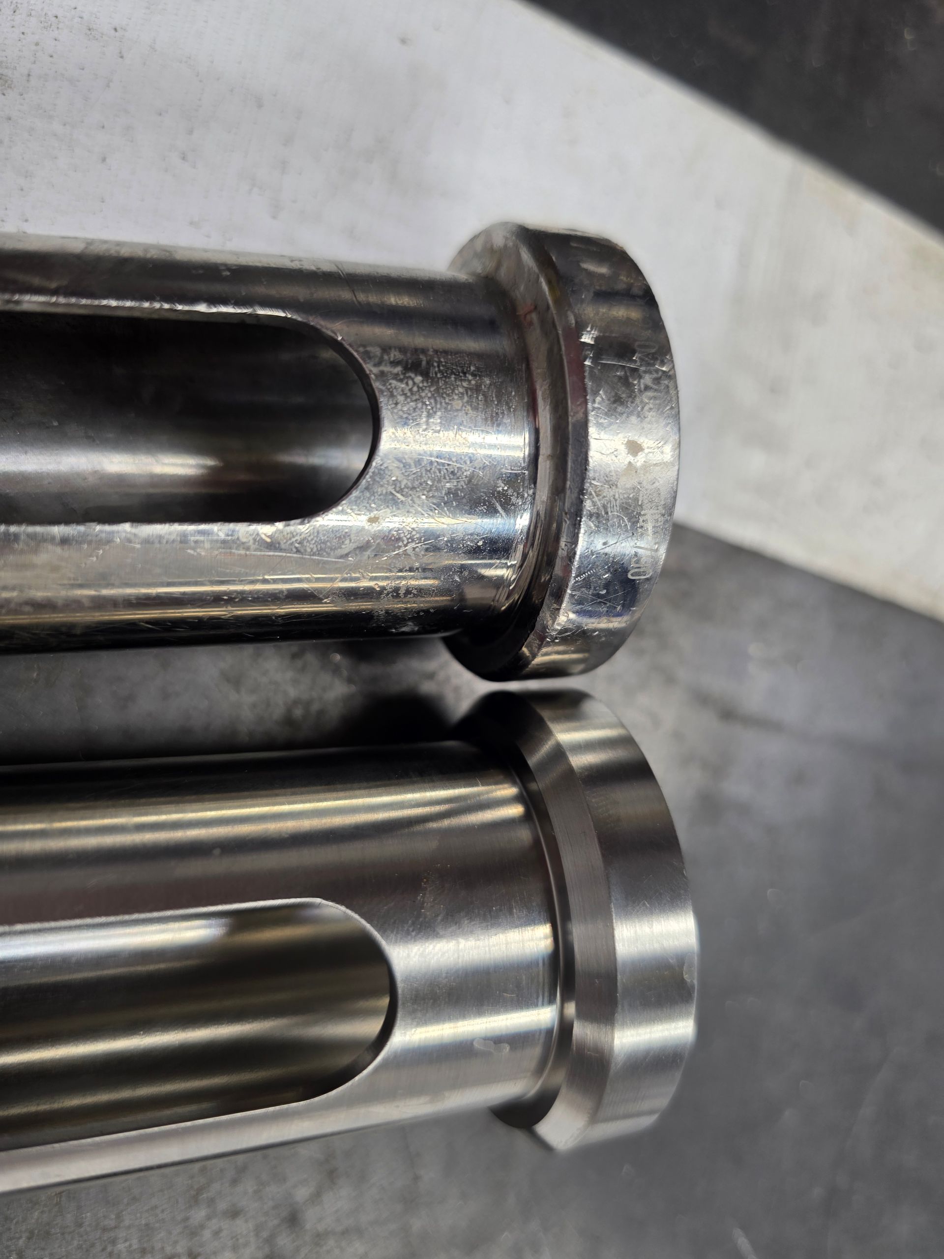 Close-up of two shiny metal slotted tubes with flanged ends on a reflective surface