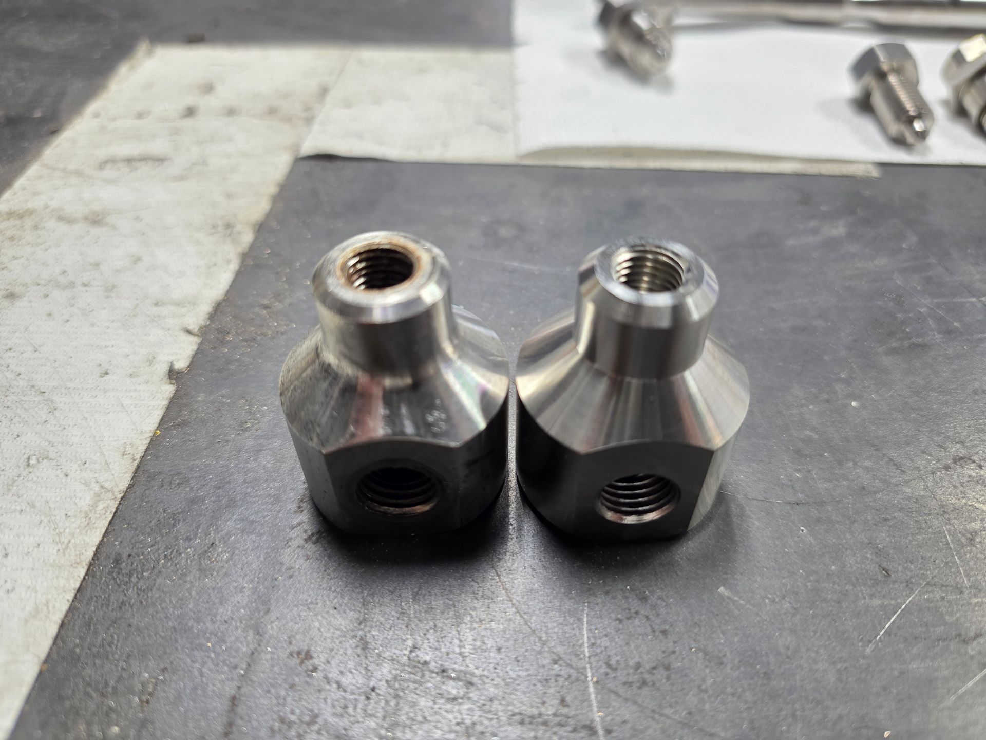 Two silver metal hex fittings on a workbench, each with threaded openings.