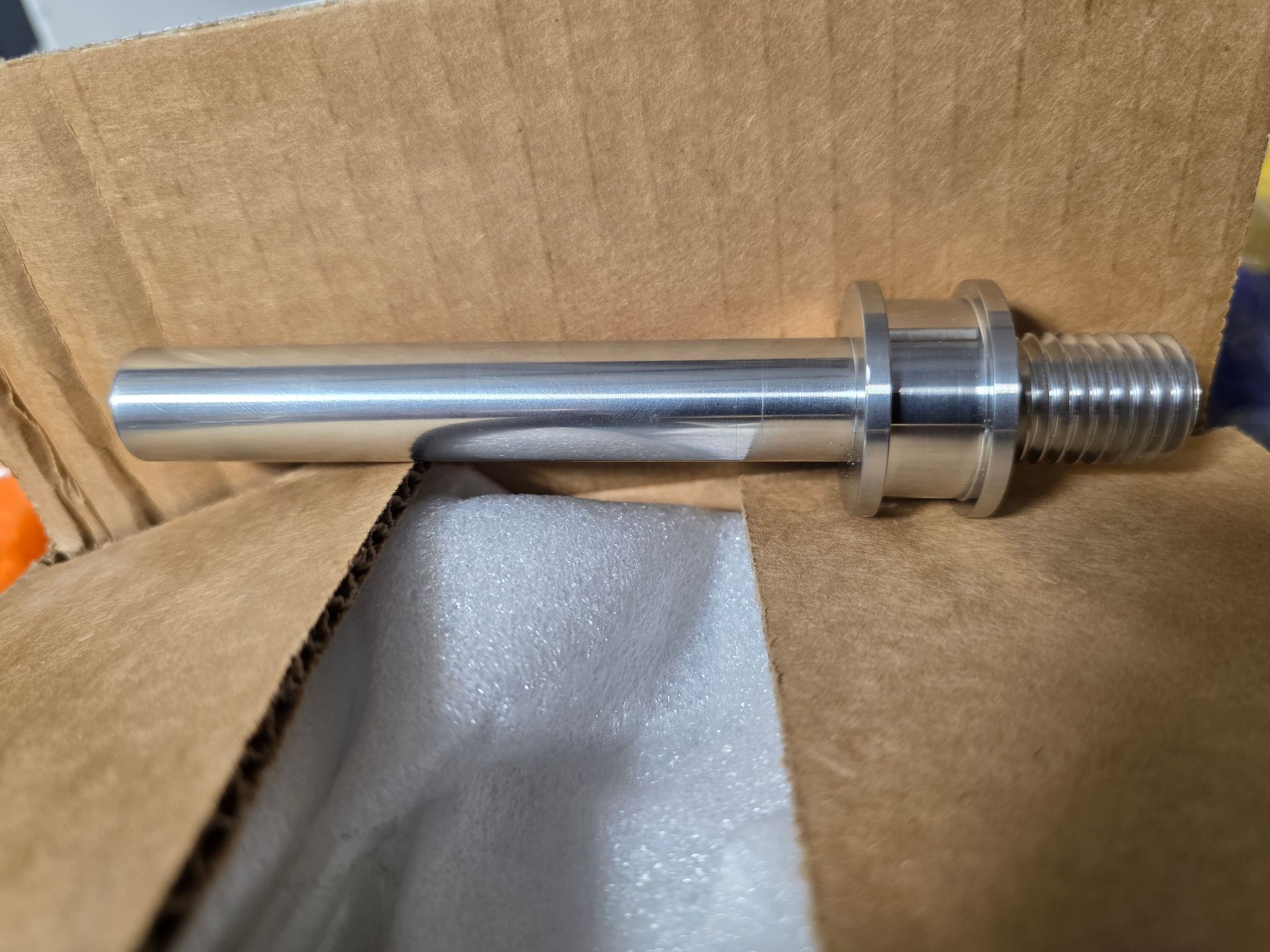 Metal cylindrical shaft with threaded end and collar, resting in a cardboard box with foam padding