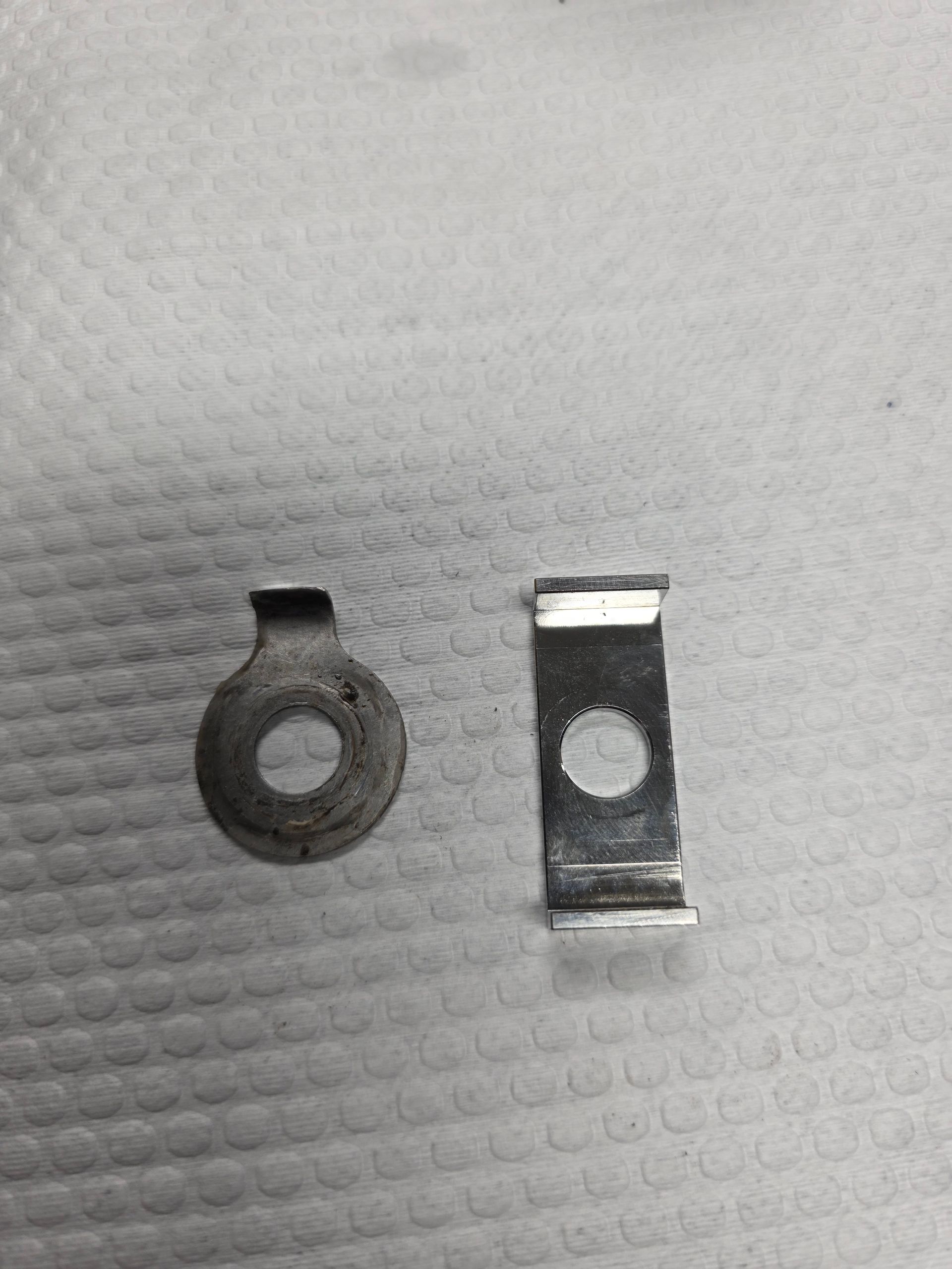 Two small metal parts on a white textured surface, one round and one rectangular with a hole.