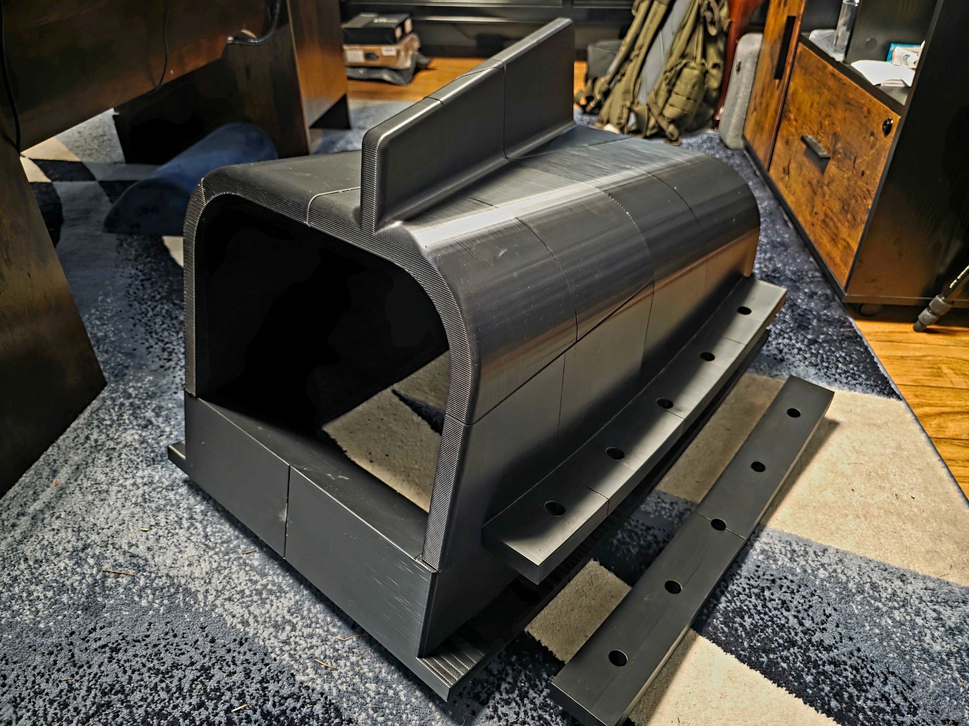 Black metal duct or hood assembly resting on a workshop floor with bolts beside it