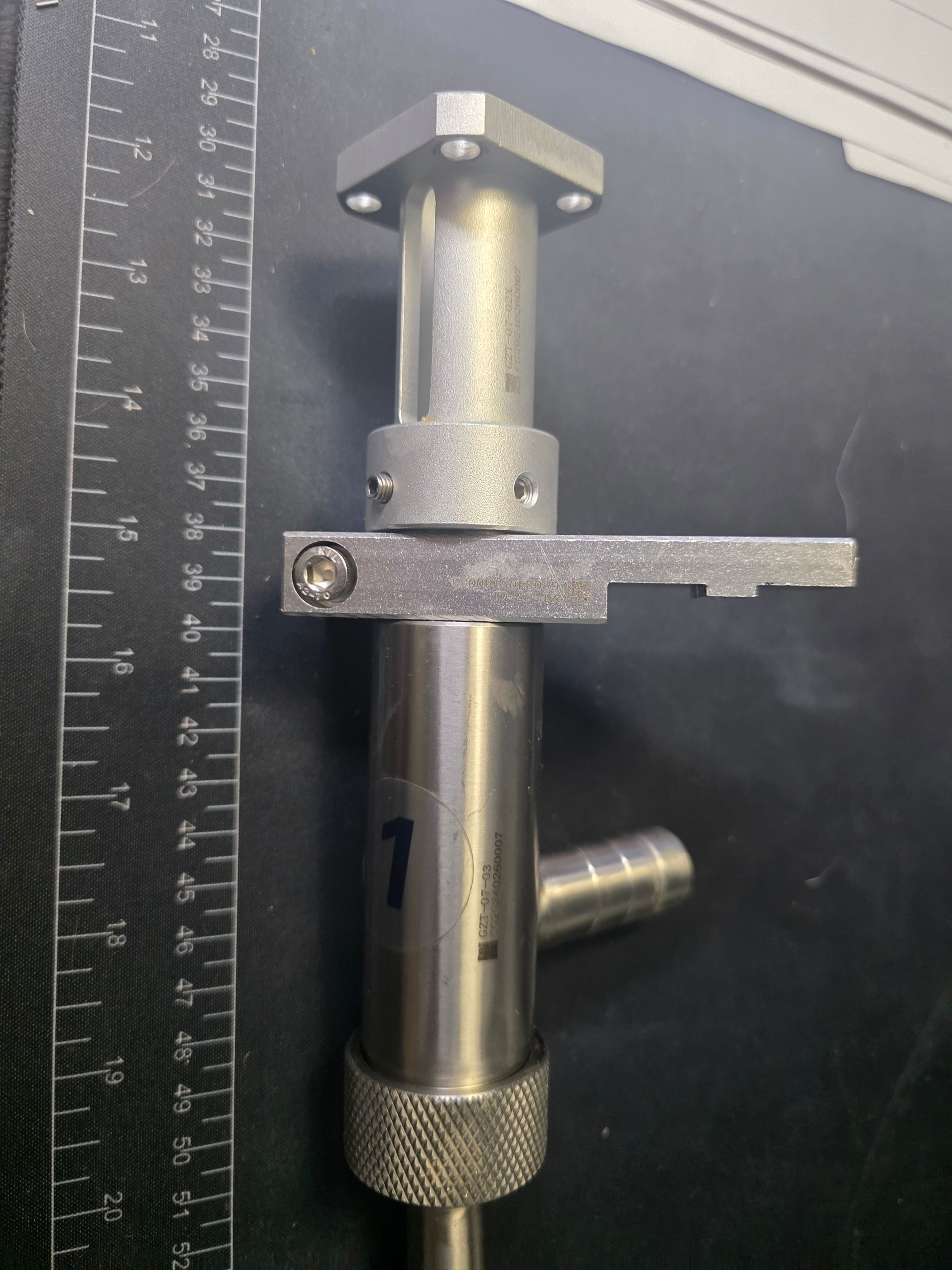Metal instrument with a ruler beside it on a dark surface, showing a horizontal arm and knurled base.