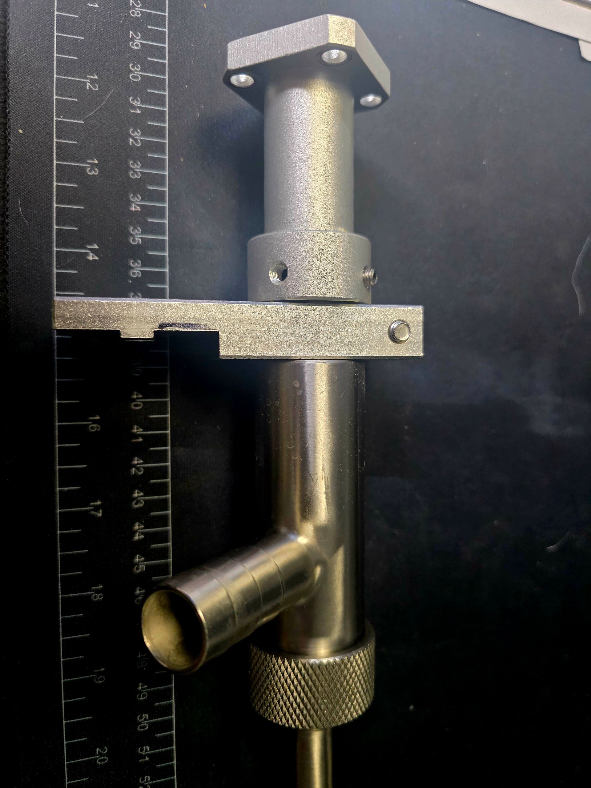 Metal pipe fitting with a bracket and knurled base against a black background, shown beside a ruler.