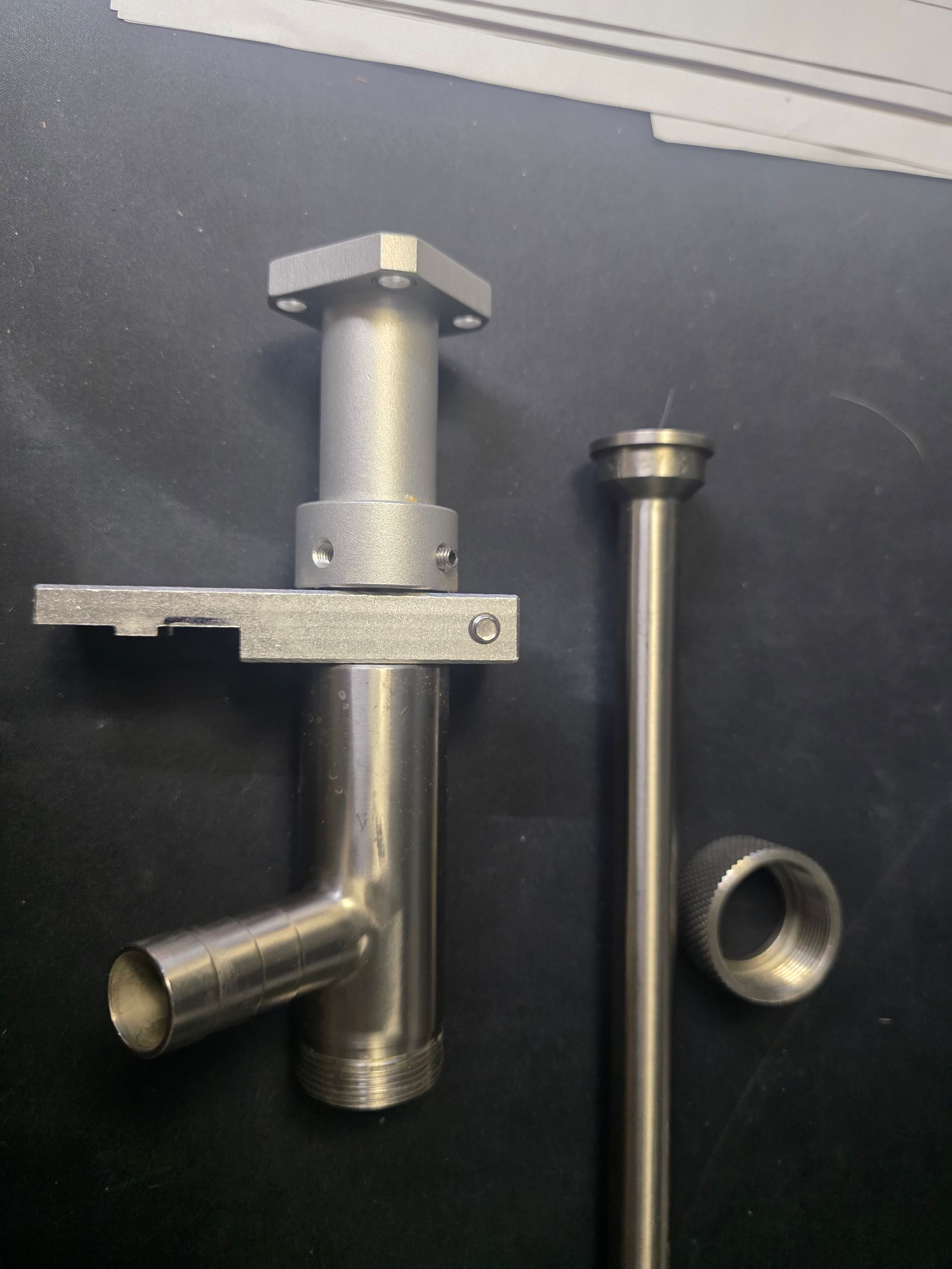 Metal pipe and fittings arranged on a dark surface, including a mounted valve-like assembly and loose parts.