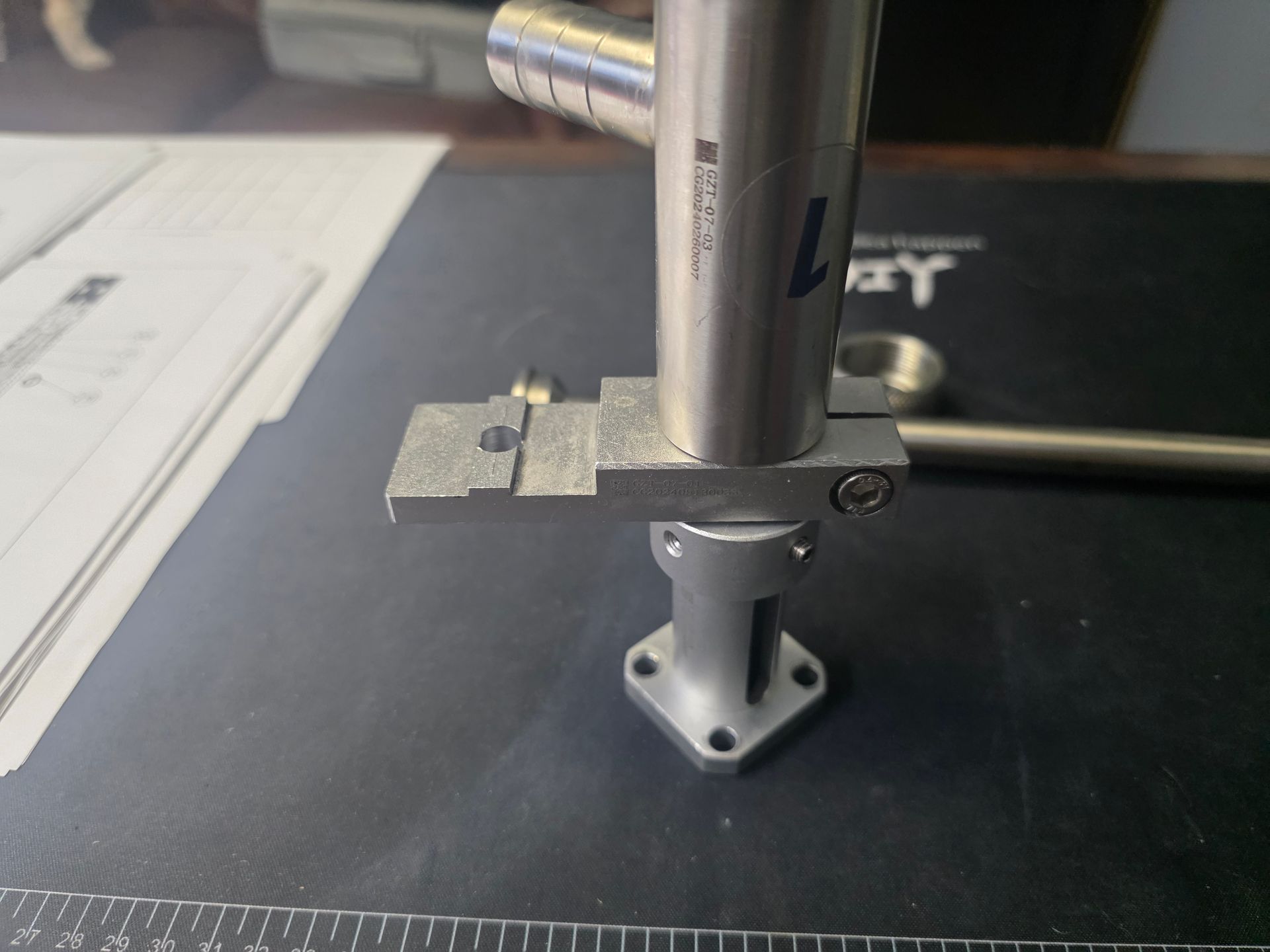 Metal clamp holding a vertical silver tube on a black workbench, with papers in the background.