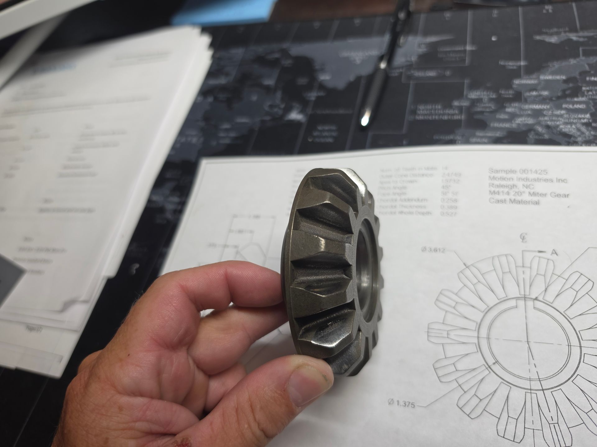 Hand holding a metal gear part over technical drawings on a desk