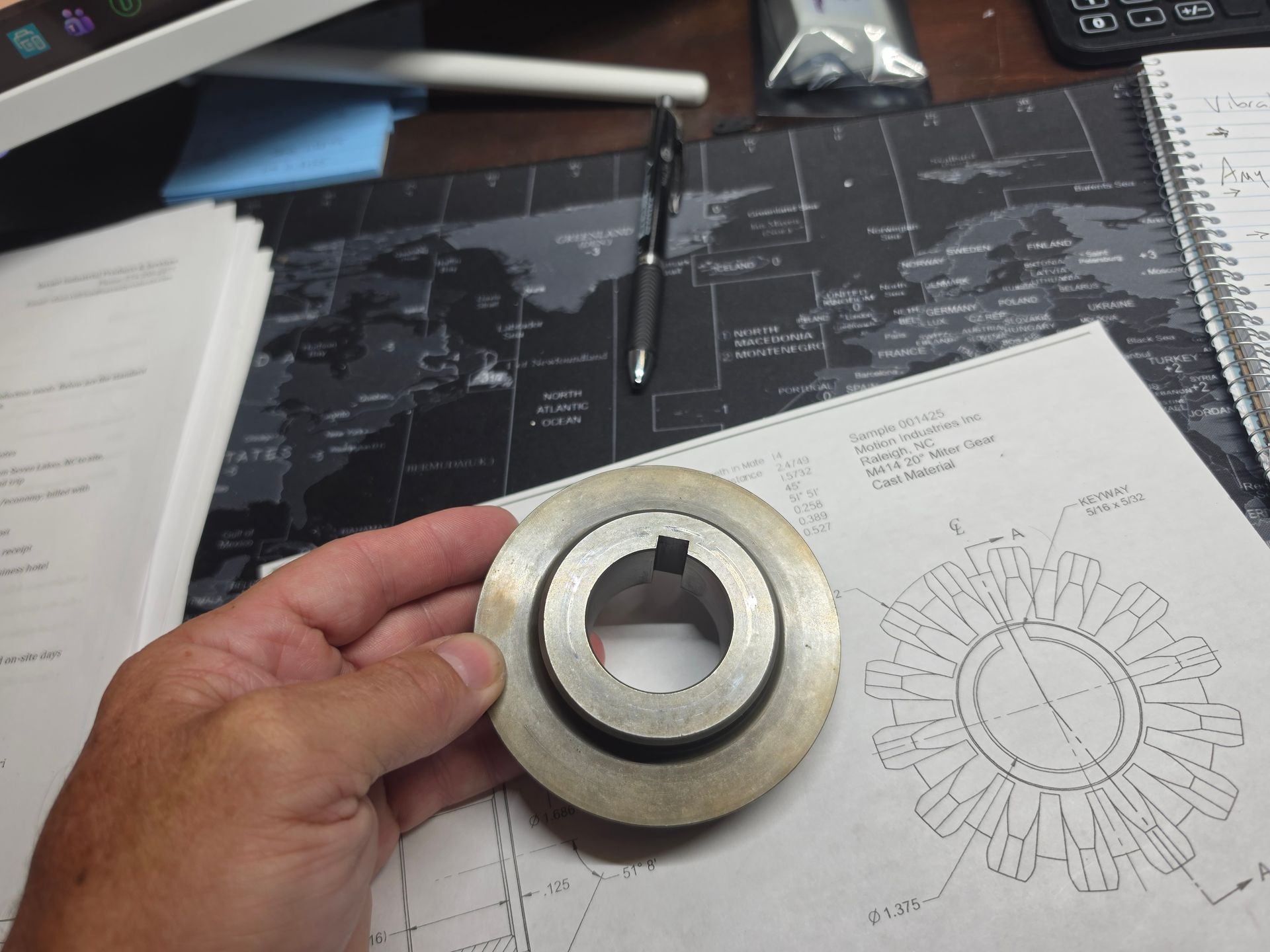 Hand holding a metal gear-like part over a desk with technical drawings and papers.