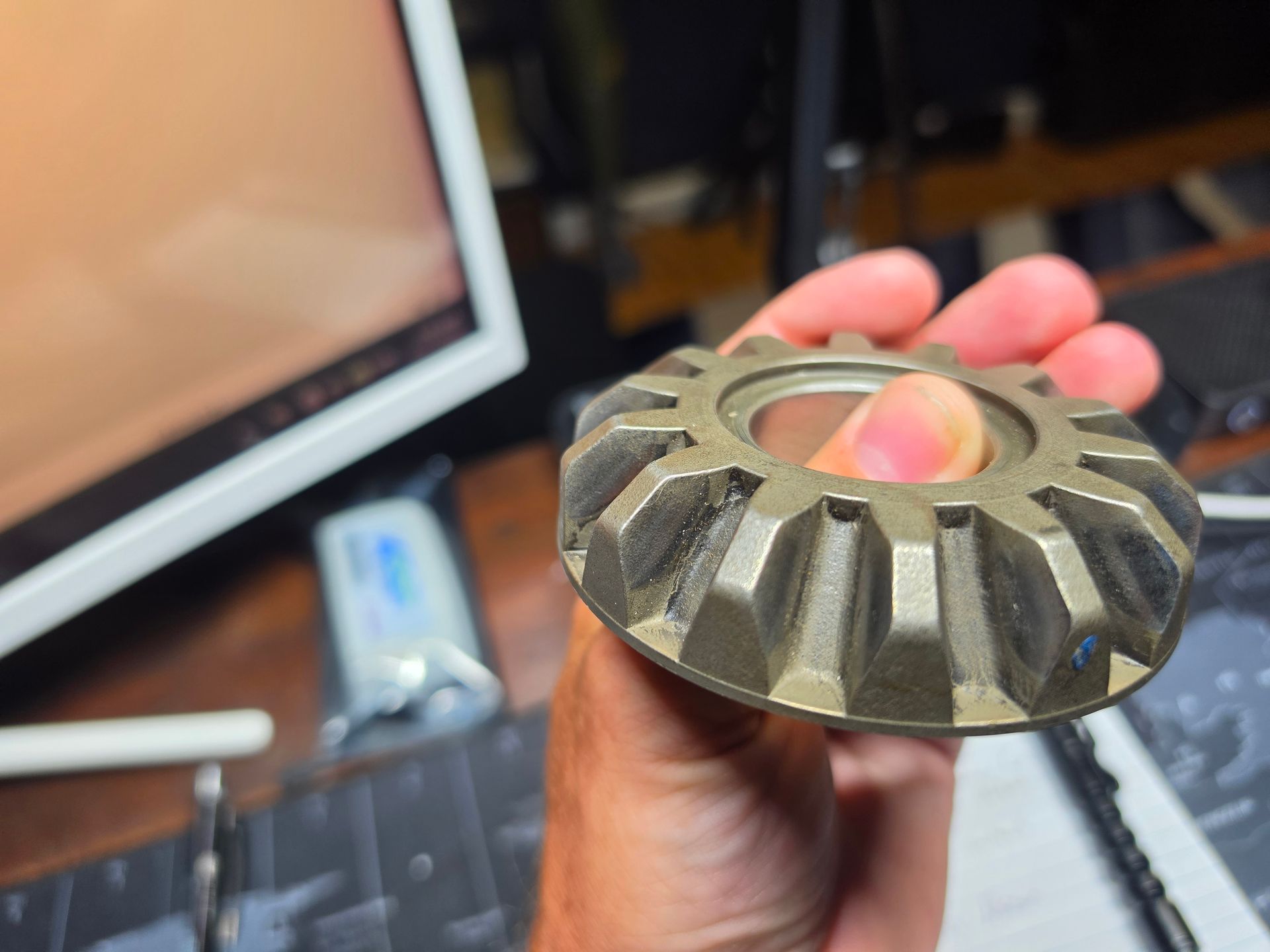 Hand holding a metal gear-like part in front of a blurred computer monitor and desk