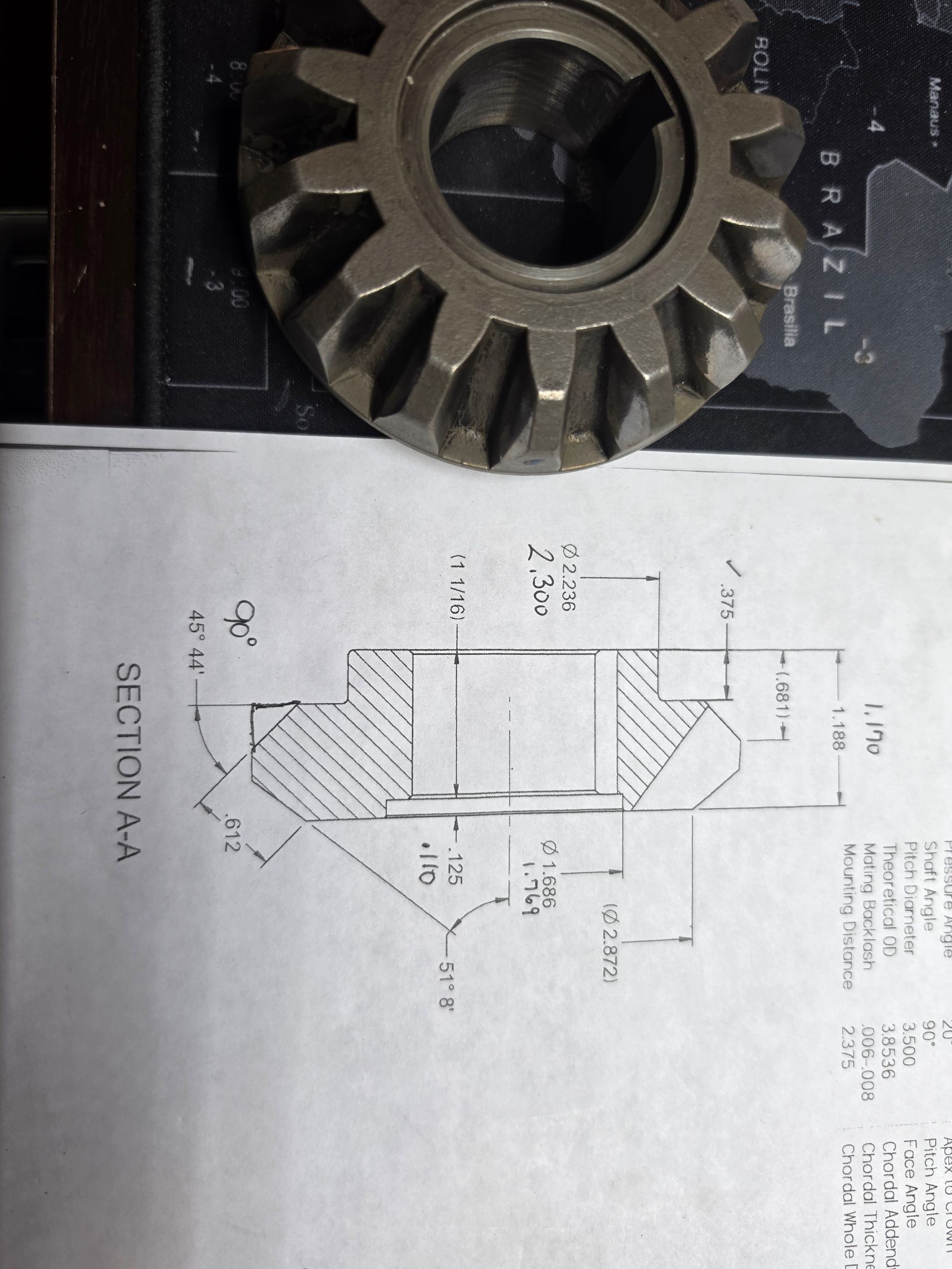 Metal gear wheel beside a technical drawing on paper, viewed from above.