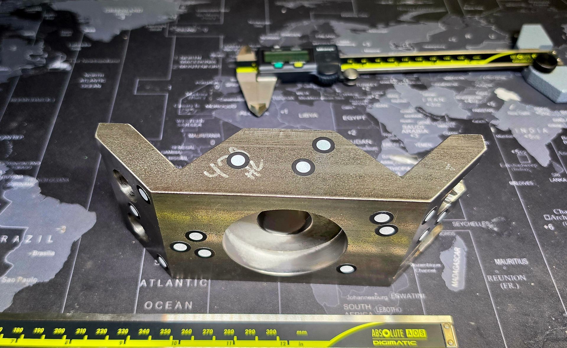 Metal bracket and caliper on a dark world-map measuring mat, with a ruler along the bottom edge.
