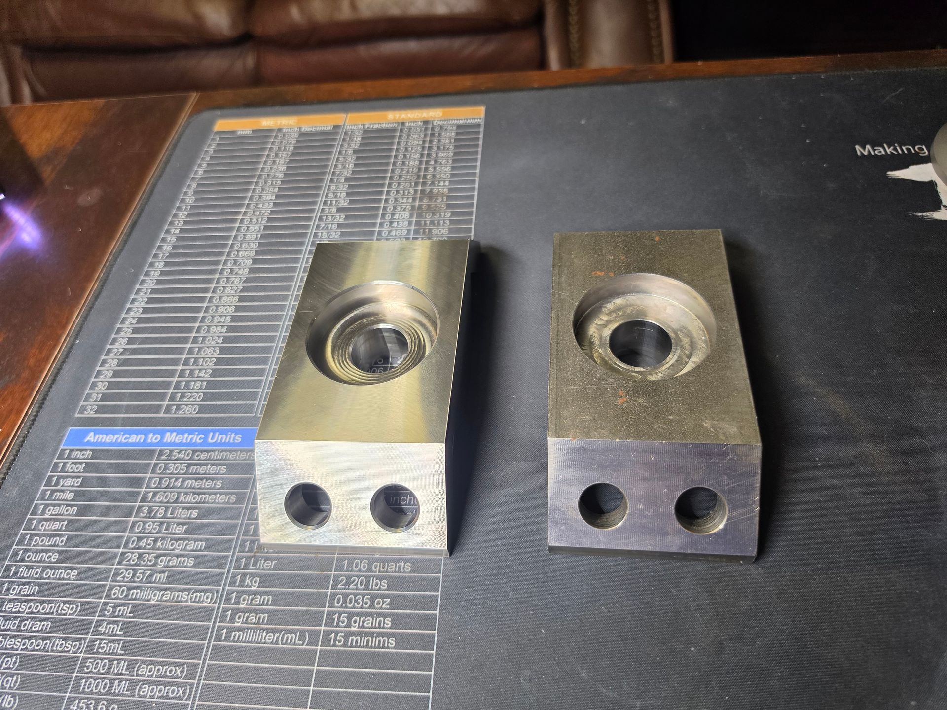 Two metal machined blocks with holes, sitting on a dark desk mat next to a ruler.