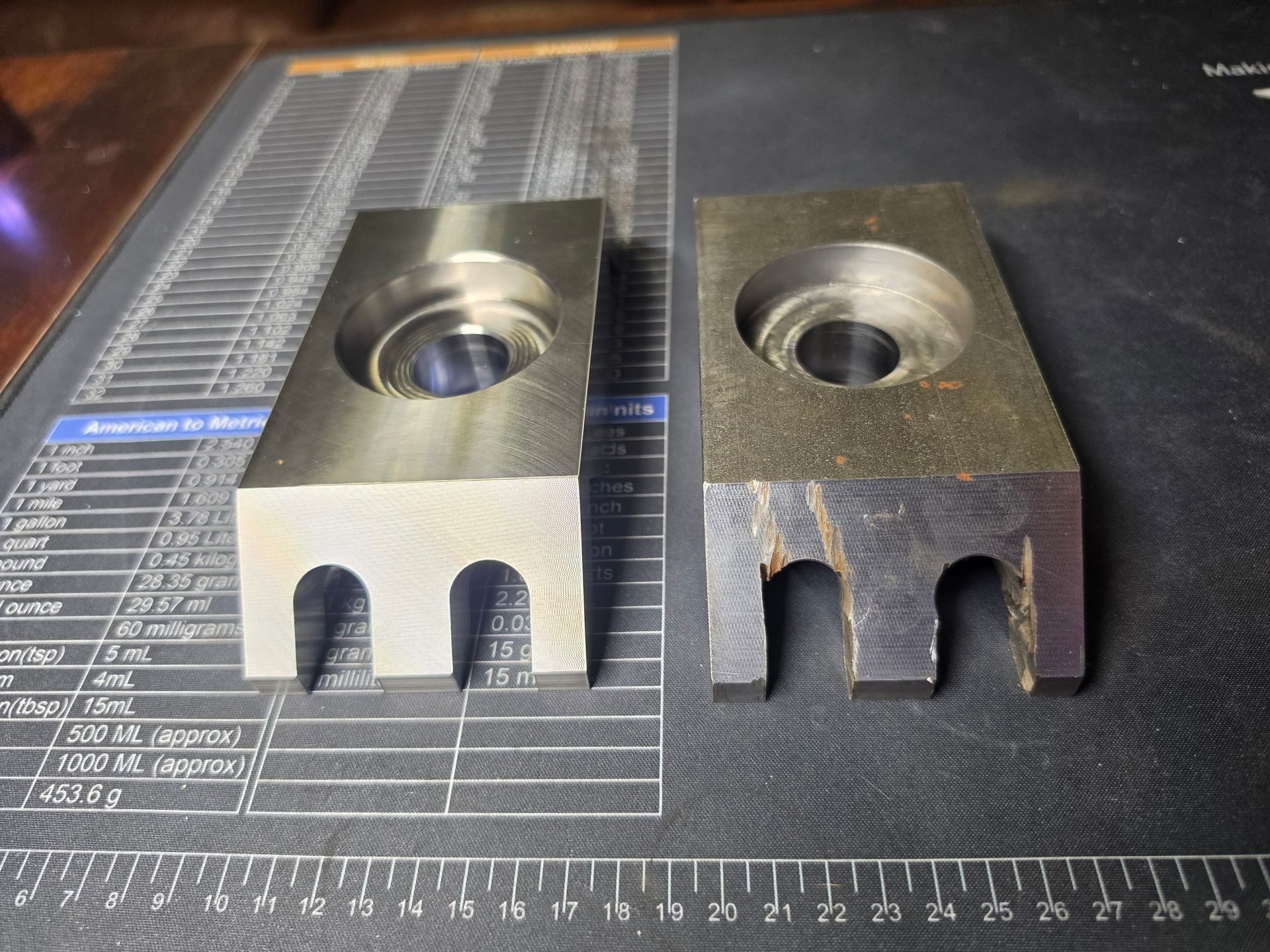 Two metal machined blocks with circular recesses and forked ends, side by side on a measurement mat.