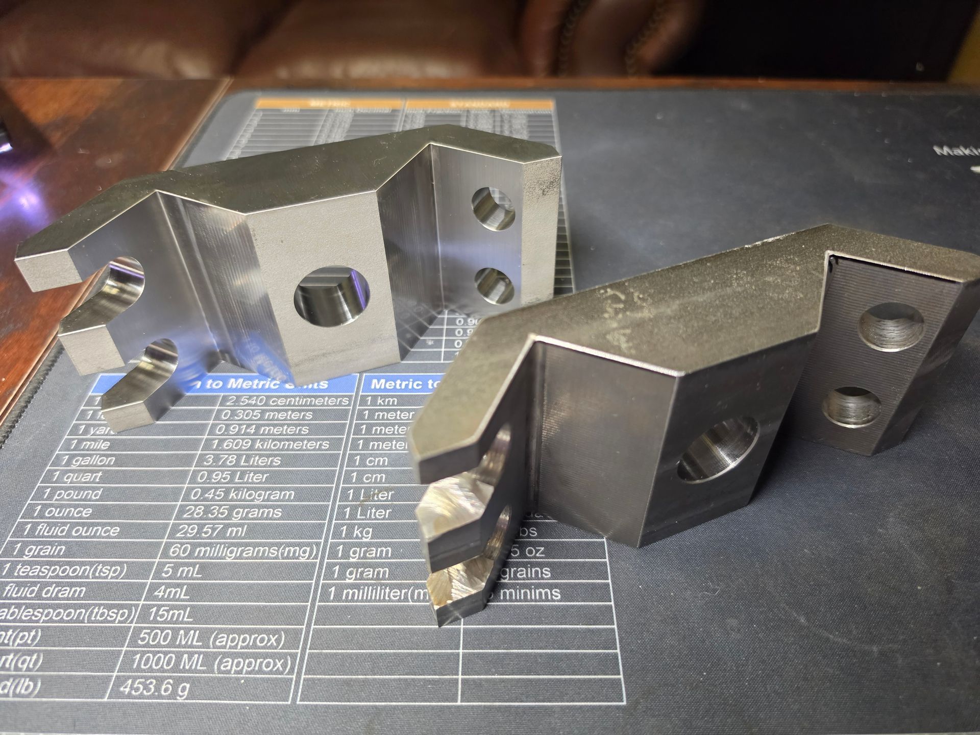 Two metal bracket parts on a printed measurement sheet, one silver and one dark gray.