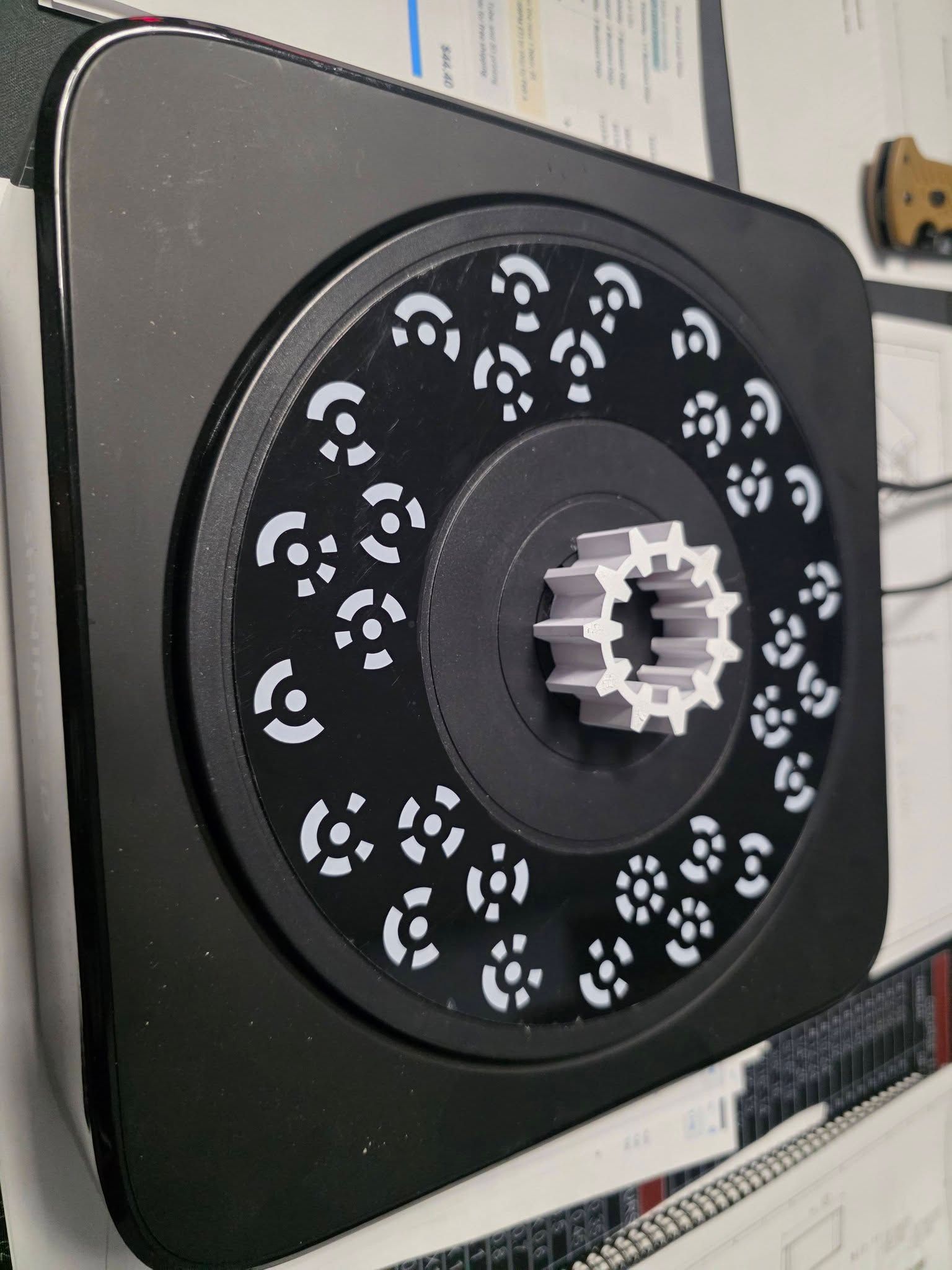 Black rotary table with white patterned lights; holds a 3D-printed gear-shaped object.