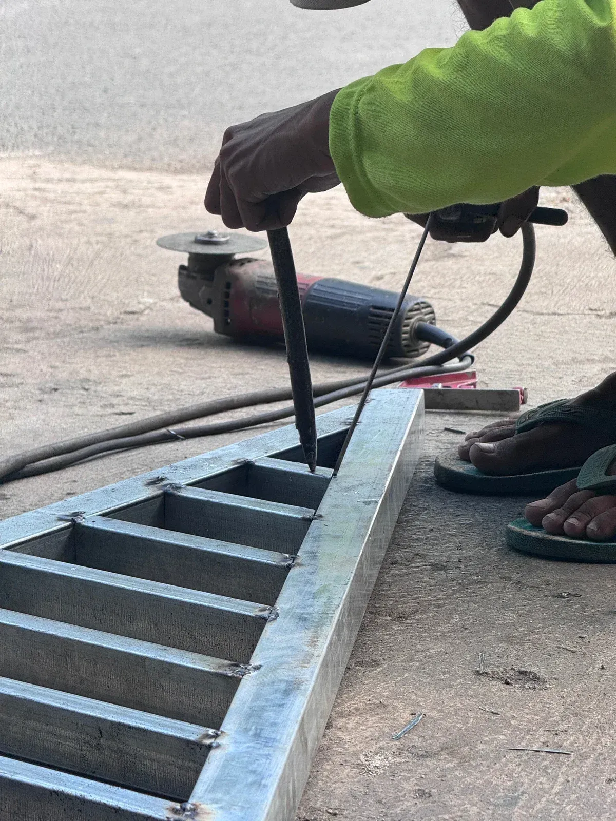 Person in green jacket lifting a metal drain grate with a tool on a sidewalk.