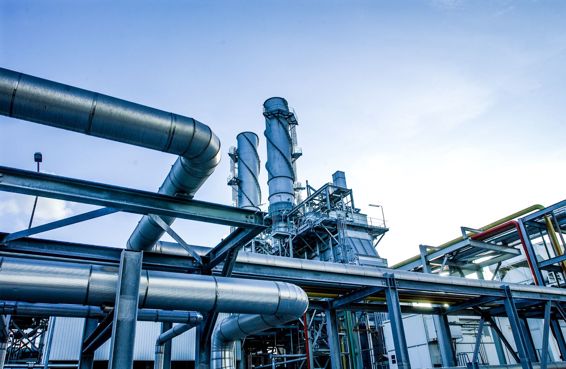 Industrial facility with large metal pipelines, exhaust stacks, and structural framework.
