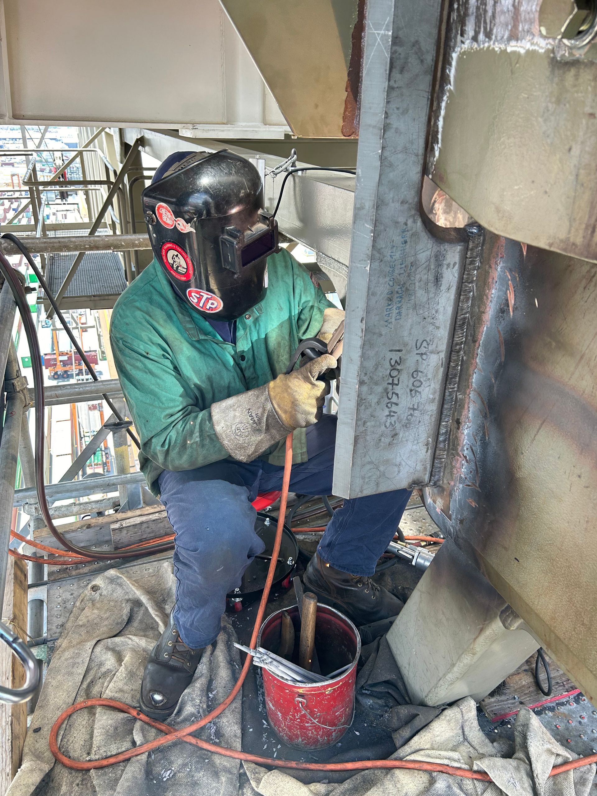 Worker in a welding mask and green jacket working with a power tool beside a metal beam.