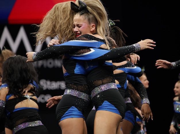 Cheerleaders in blue and black uniforms embracing, some crying, on a competition stage.