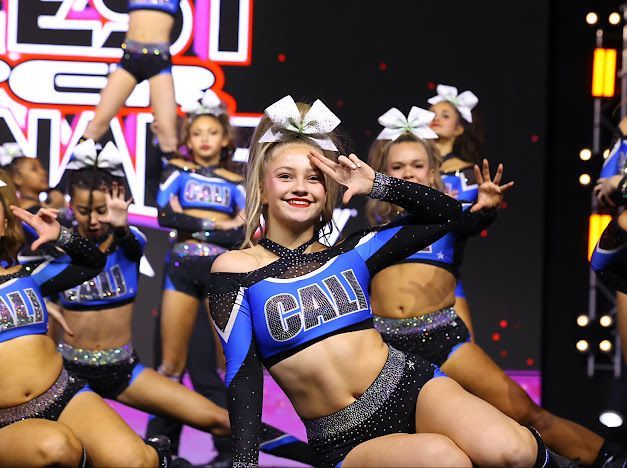 Cheerleaders in blue and black uniforms performing. A girl smiles, making a salute, at a competition.
