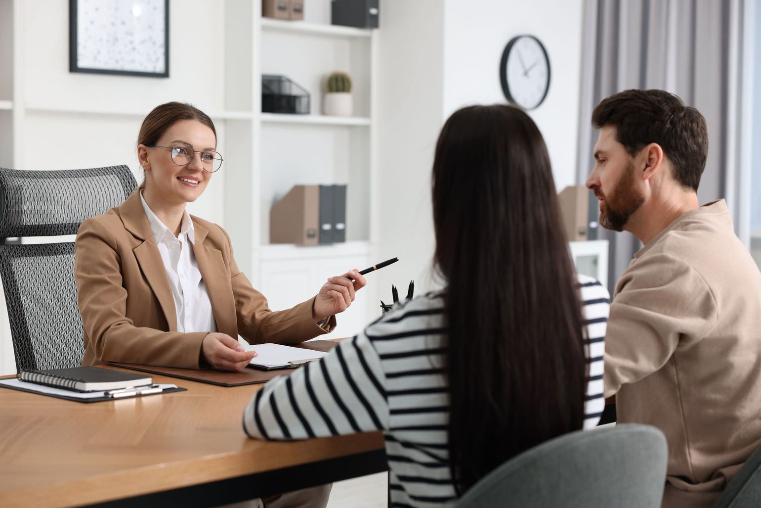 A professional, smiling person in a suit points to a document while consulting with a couple at a desk.