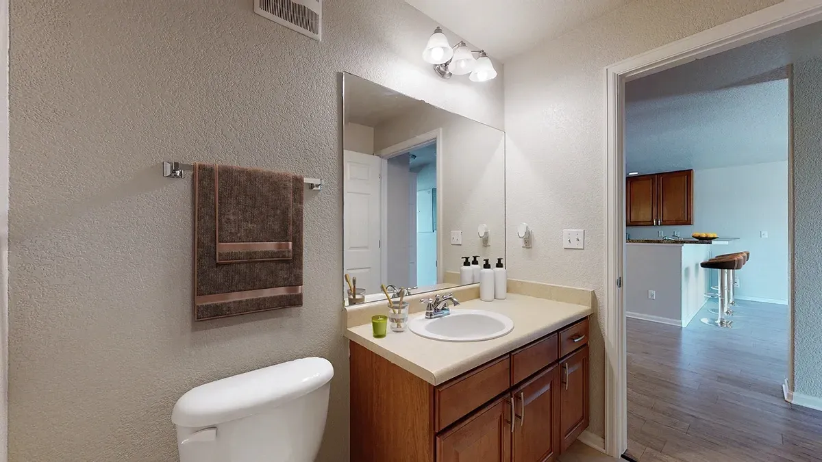 Bathroom vanity with sink, mirror, and toilet, leading to a kitchen area.