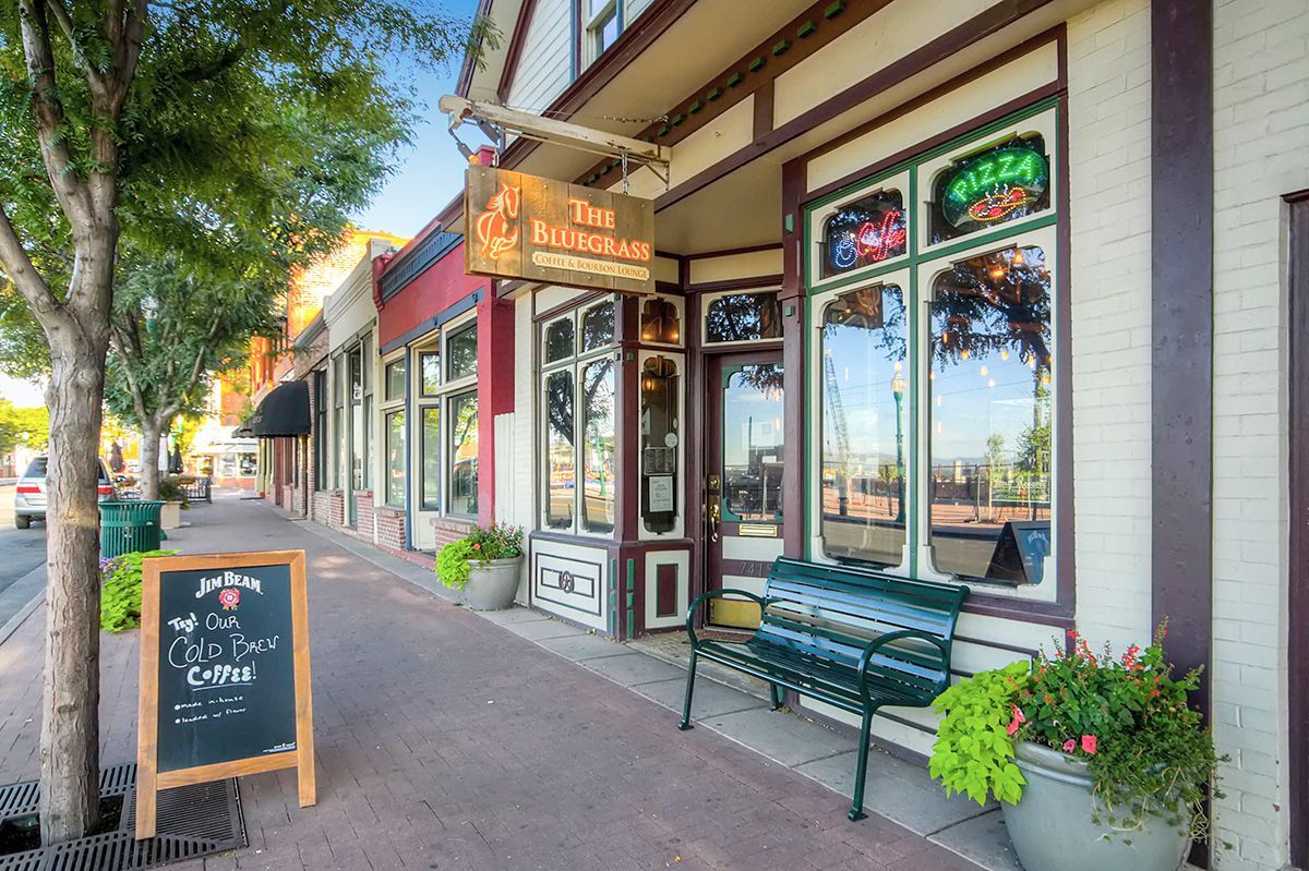 Small-town storefronts with a chalkboard sign, green bench, and flower planters along a sunny sidewalk