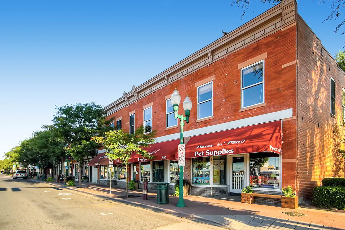 Brick storefronts with red awnings on a sunny small-town street, with trees and parked cars nearby