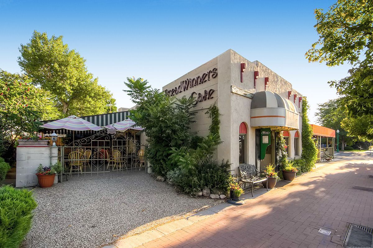 Sunny outdoor cafe with a gravel patio, umbrellas, greenery, and a white stucco building on a quiet street