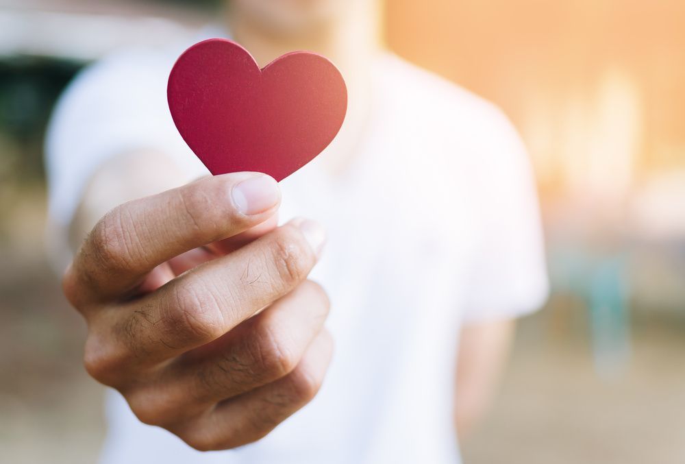 Hand holding a small red heart-shaped cutout against a blurred bright background