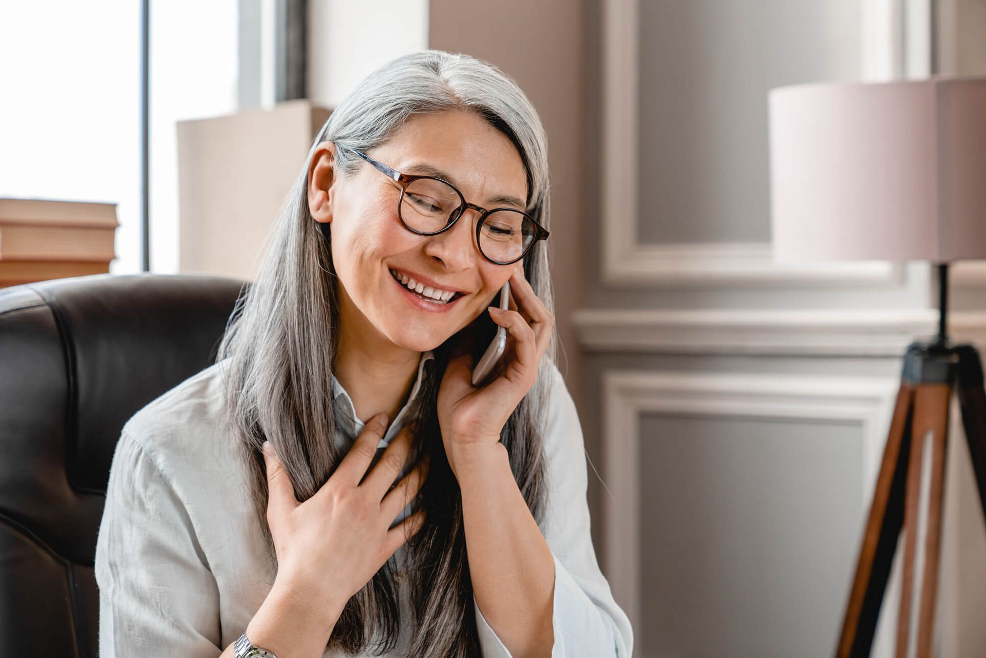 Smiling person in glasses and a gray sweater talking on a phone in a bright room