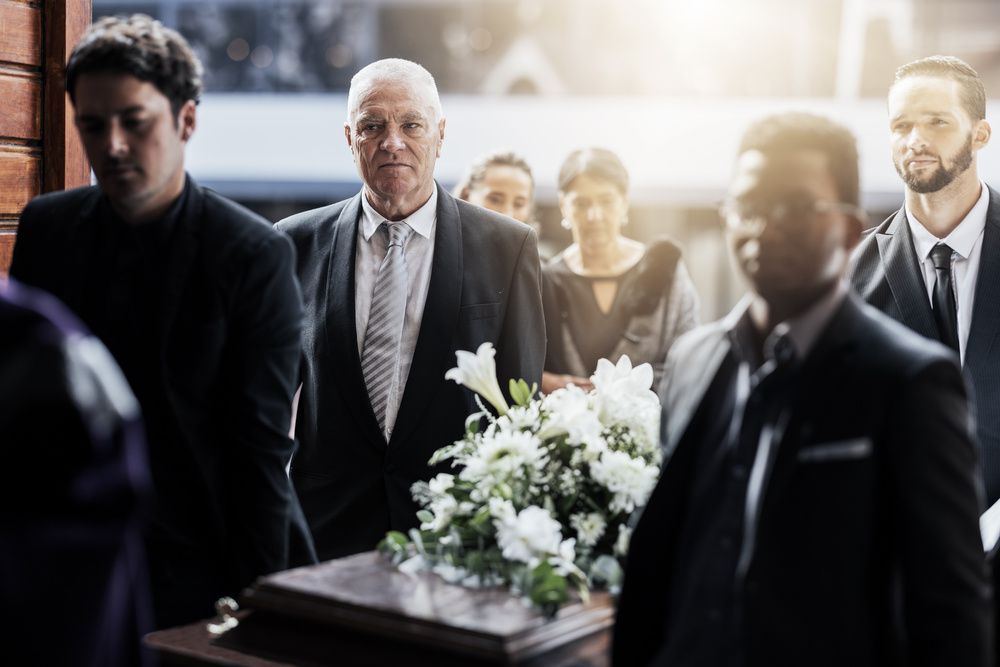 Mourners stand around a casket adorned with white flowers at a funeral service.