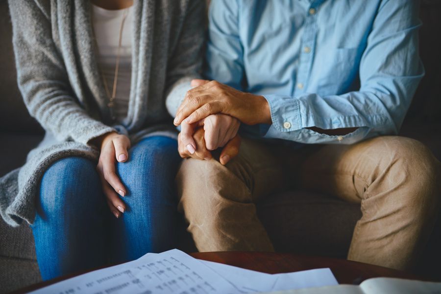 Two people sitting on a couch, holding hands over their knees, with papers on the table in front.