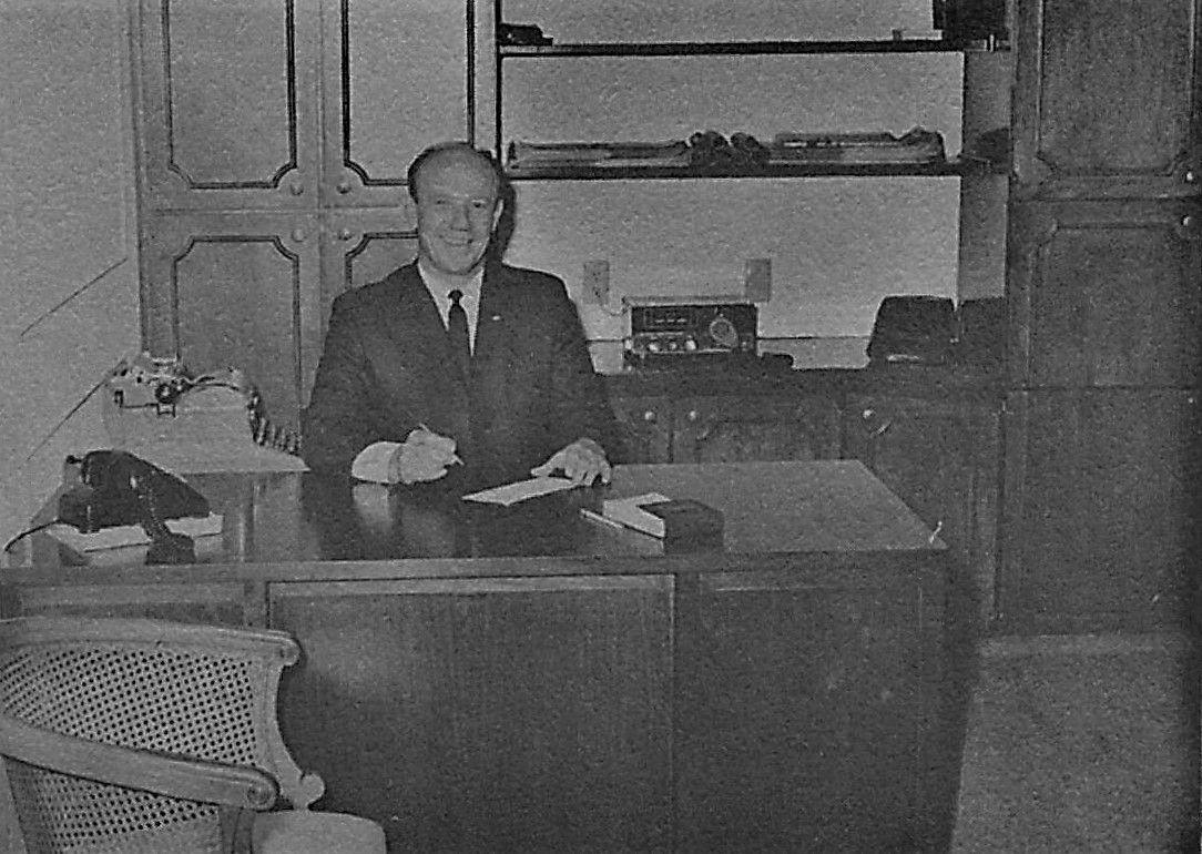 Man seated at a desk in an office, holding papers in a black-and-white scene