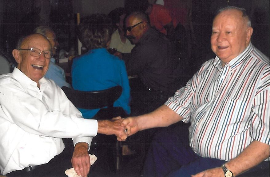 Two smiling men seated indoors shaking hands, one in a white shirt and one in a striped shirt.