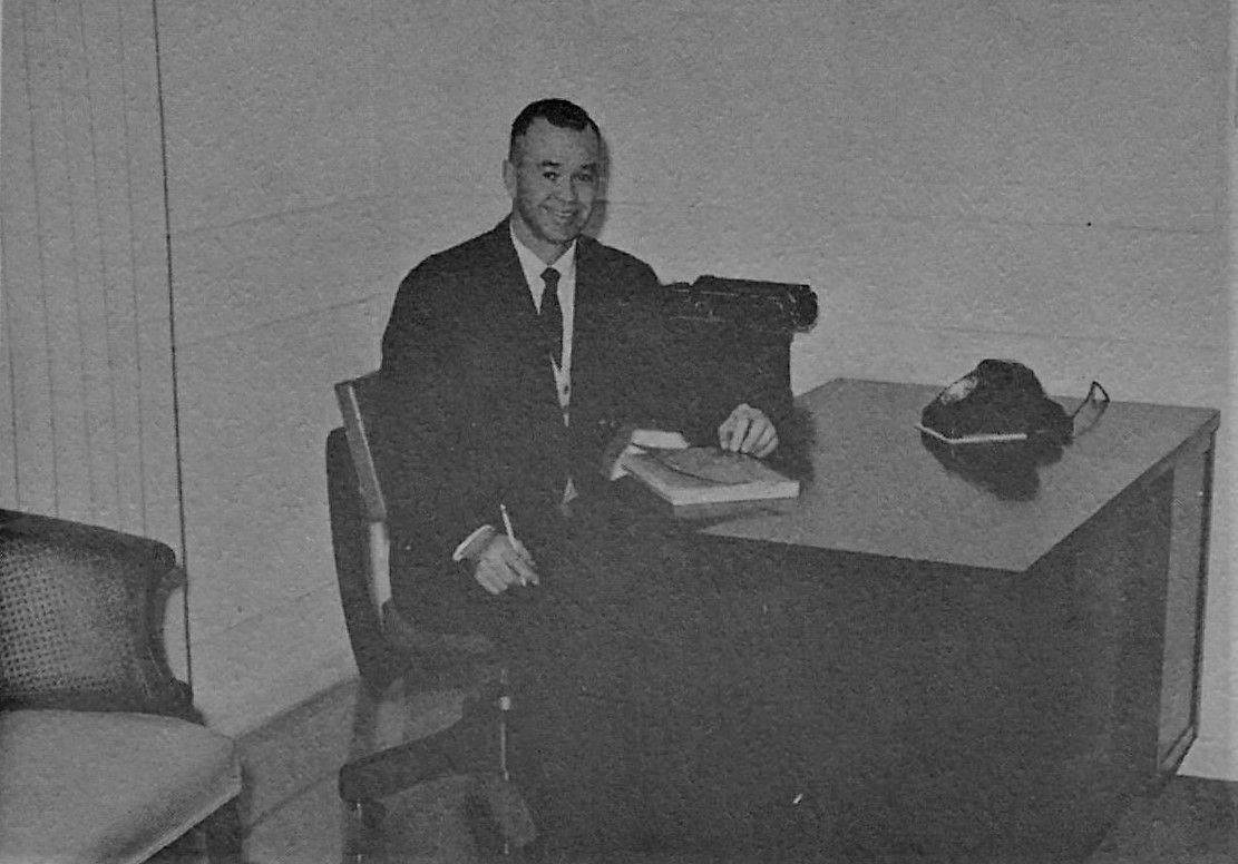 Man in a suit seated at a desk in a sparse office, with a telephone and papers nearby.